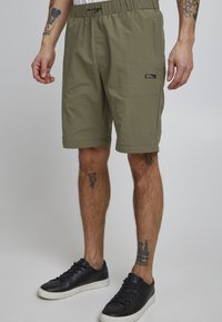 Olive green shorts with elastic waistband, drawstring, and side pocket. Made of lightweight fabric with a smooth texture. Black label detail.