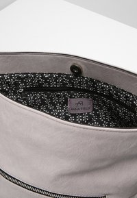 Grå läderväska insida med ett blommönstrat svart tygfoder och en synlig dragkedjeficka, med en etikett som läser "ANNA FIELD."