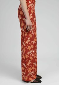 Pantalons rouges à motifs présentant un design floral et animal en orange. Tissu léger avec une coupe ample, associés à des sandales noires.