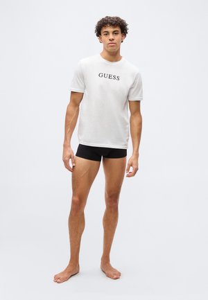 Giovane uomo con capelli ricci in piedi a piedi nudi, indossa una t-shirt bianca Guess e pantaloncini aderenti neri su uno sfondo chiaro e uniforme.