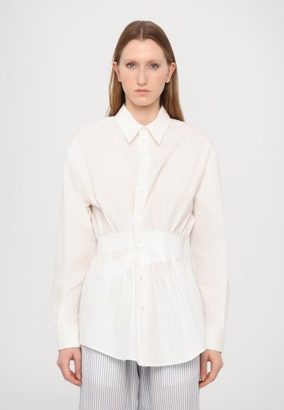 Chemise blanche à boutons avec une taille cintrée, dotée d'une ceinture transparente, associée à un pantalon rayé, en tissu lisse, et à manches longues.