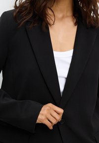 Blazer noir avec un col à revers entaillé, fermeture à un seul bouton et texture lisse, porté sur un débardeur blanc côtelé.