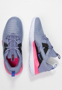 Zapatillas deportivas de color gris claro con una parte superior texturizada, acentos negros, suelas rosas y detalles en azul y rosa en el talón y la lengüeta.