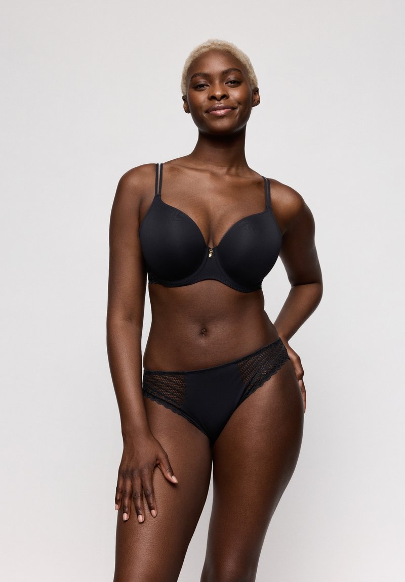 Vrouw met kort blond haar, dragend een zwarte beha en bijpassende slip met kanten rand, staand tegen een eenvoudige lichte achtergrond.