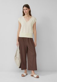 Jasno beżowy sweter z dekoltem w serek połączony z szerokimi brązowymi spodniami. Modelka trzyma kremową torebkę z koralików i nosi białe sandały typu slide. Widoczne gładkie tekstury.