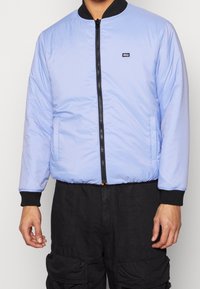 Veste bomber bleu clair avec un col côtelé noir, une fermeture éclair à l'avant et des poches latérales. Présente un petit écusson logo sur la poitrine gauche.
