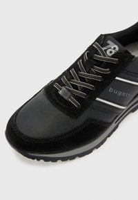 Chaussure de sport noire avec une tige en cuir lisse et en daim. Elle est dotée de lacets gris, de rayures blanches horizontales et d'une semelle en caoutchouc texturée.