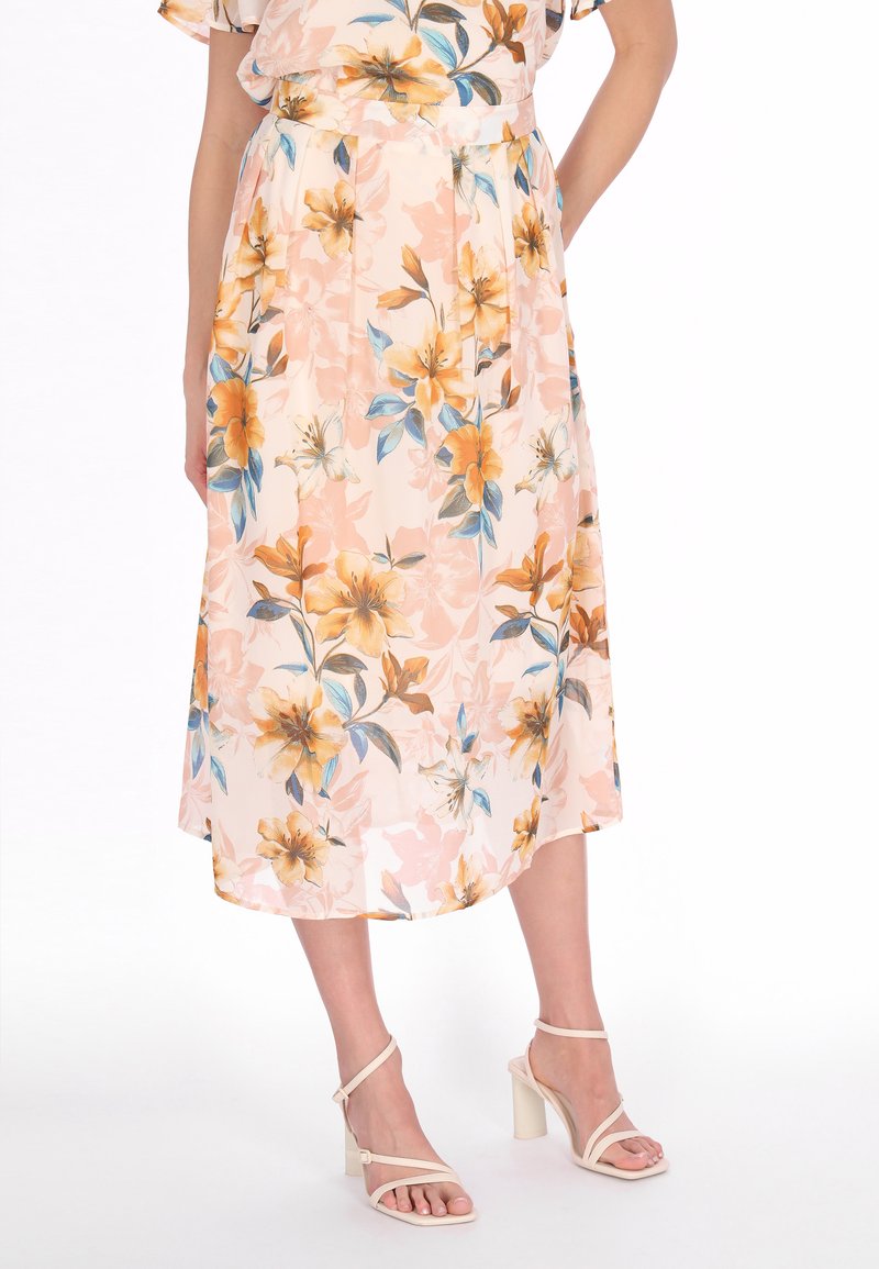 Frau trägt einen pfirsichfarbenen Midirock mit Blumenmuster und nudefarbene Riemchen-Sandaletten mit Blockabsatz, steht mit einer Hand in der Hosentasche.