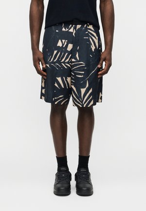 Άνδρας φοράει μαύρα και μπεζ σορτς με αφηρημένο σχέδιο φύλλων, μαύρες κάλτσες και μαύρα αθλητικά παπούτσια, στέκεται μπροστά σε απλό ανοιχτό φόντο.