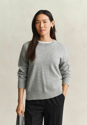 Vrouw met lang donker haar, gekleed in een grijze trui en zwarte broek, staand met één hand in haar zak en met de andere hand stof vasthoudend.