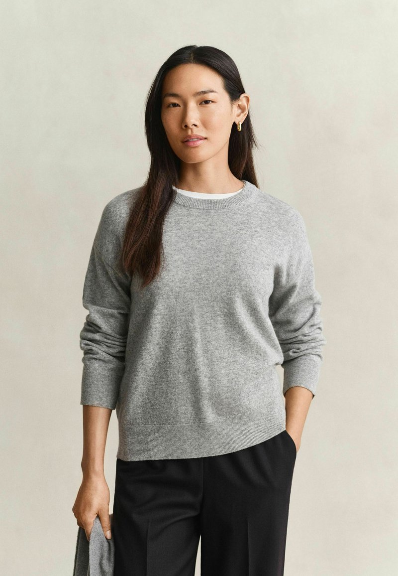 Femme aux longs cheveux foncés portant un pull gris et un pantalon noir, debout, une main dans la poche et tenant un tissu dans l'autre main.