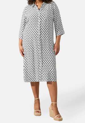 Frau trägt ein knielanges weißes Kleid mit schwarzem Rautenmuster, dreiviertelärmeln, Vorderknöpfen und beigen Keilsandalen.