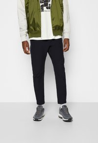 Giacca bomber verde sopra una t-shirt bianca con grafica, abbinata a pantaloni slim-fit blu navy e sneakers grigie con dettagli testurizzati e lacci.