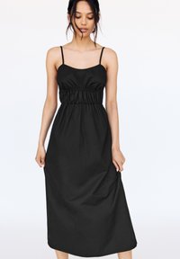 Robe maxi noire avec des fines bretelles, taille froncée et tissu lisse légèrement texturé. Design simple et coupe longue.