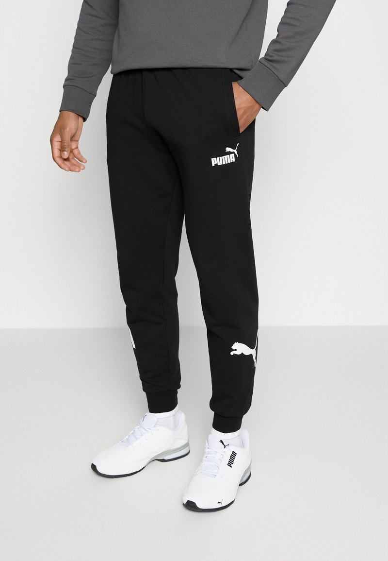 Černé tepláky s elastickým pasem, logo PUMA v bílém na levém stehně a ze stran. Nošeny s bílými sportovními botami. Hladký bavlněný materiál.