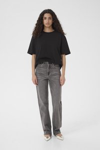 Kobieta o kręconych włosach, ubrana w czarny, za duży t-shirt, szare jeansy z wysokim stanem oraz białe, spiczaste buty z metalicznym detalem.