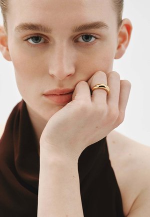 Anillo de oro con un diseño suave y redondeado, llevado en una mano que descansa sobre una barbilla. El tono de piel es claro; el fondo es blanco, con detalles mínimos de la ropa.