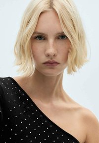 Top negru cu umeri goi, decorat cu puncte argintii. Modelul are părul scurt, ondulat, blond și un fundal neutru.