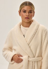 Kremowy szlafrok o miękkiej, teksturowanej tkaninie, z kołnierzem szalowym, paskiem w talii i puszystym, przytulnym wykończeniem.