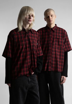 Dvaja ľudia oblečení v červených a čierno-károvaných krátkych rukávoch cez čierne tričká s dlhým rukávom, stojaci pred jednoduchým sivým pozadím.