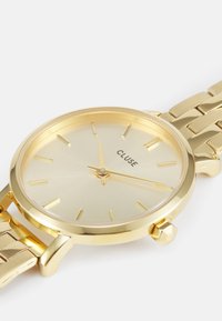 Gold metal wristwatch with a minimalist round face, sunburst texture, gold hour markers, and a linked bracelet strap. Brand name "CLUSE" displayed.