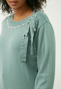Maglione turchese chiaro con maniche lunghe, presenta un collo decorativo di perle e un accent con fiocco impreziosito da perline sparse.