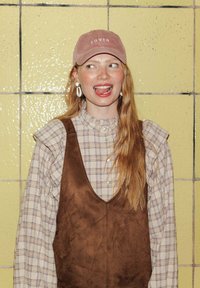 Salopette en daim marron sur une chemise à carreaux, avec un col volant, associée à une casquette en velours côtelé rose étiquetée "LOVER" et des boucles d'oreilles blanches.