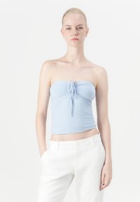 Top de culoare albastru deschis, fără bretele, cu un detaliu de legare în față, realizat dintr-un material neted; purtat cu pantaloni albi, lejeri.