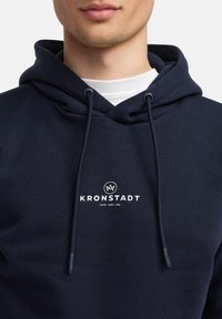 Hoodie bleu marine avec un cordon, arborant un logo et un texte blancs sur la poitrine. Fabriqué en tissu doux avec une surface texturée.