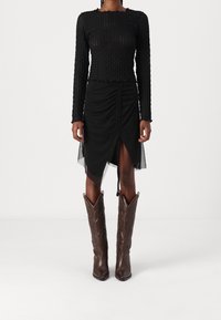 Svart texturerad långärmad top som matchas med en svart rynkad kjol med ett mesh-overlay och en hög springa, kompletterad med bruna cowboystövlar.