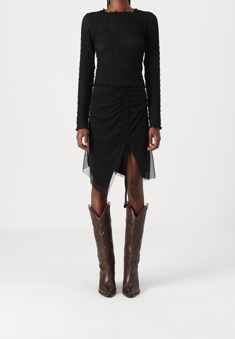 Svart texturerad långärmad top som matchas med en svart rynkad kjol med ett mesh-overlay och en hög springa, kompletterad med bruna cowboystövlar.