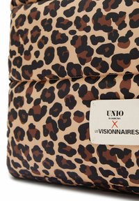 Leopardenmusterstoff mit einem aufgenähten weißen Etikett, auf dem in schwarzem und rotem Text "UNIO Hamburg X Les Visionnaires" steht.