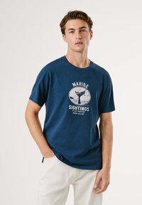 Camiseta de algodón de color azul marino con mangas cortas, escote redondo y un estampado gráfico de una ballena que incluye texto. Combinada con pantalones de color claro.