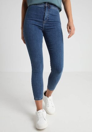 Kobieta ubrana w niebieskie, dopasowane spodnie z wysokim stanem i białe trampki, stojąca na gładkiej jasnoszarej podłodze na tle jasnoszarej ściany.