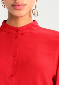 Chemise rouge boutonnée avec un col, fabriquée en tissu lisse, avec des bords arrondis et des boutons rouges assortis sur le devant.