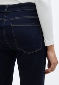 Σκούρο μπλε τζιν παντελόνι με εφαρμοστή γραμμή, που διαθέτει κίτρινη στοίχιση σε αντίθεση, δύο πίσω τσέπες και λεία υφή.