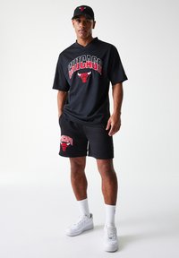 Camisola desportiva em rede preta com o logótipo vermelho dos "Chicago Bulls", combinada com calções pretos que apresentam o mesmo logótipo. Ténis e meias brancas.