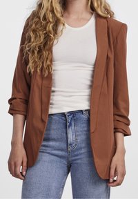 Blazer de couleur rouille avec une coupe slim, un tissu doux et des épaules structurées. Comprend des manches retroussées et des poches latérales, porté sur un haut blanc.