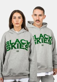 Hoodie harmaa, jossa on suuri vihreä teksti "MUSTAT TAIVAAAT" ja simpukan grafiikka, etutasku ja ribatut hihansuut.