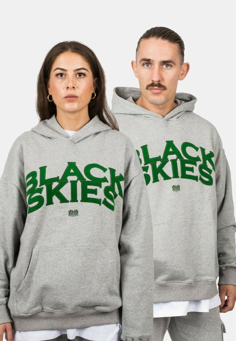 Hoodie harmaa, jossa on suuri vihreä teksti "MUSTAT TAIVAAAT" ja simpukan grafiikka, etutasku ja ribatut hihansuut.