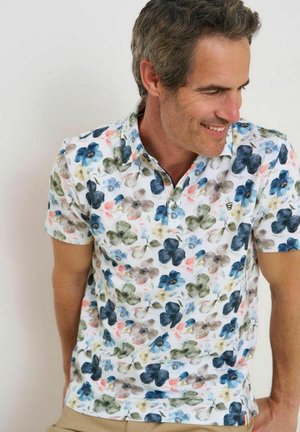 Man glimlachend, draagt een wit poloshirt met korte mouwen met een blauw, groen en roze bloemenpatroon, staand tegen een effen achtergrond.