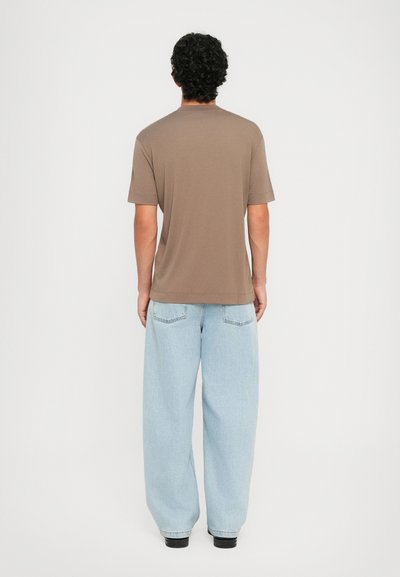 Homme aux cheveux bouclés et foncés, portant une chemise marron à manches courtes et un jean léger bleu clair, se tenant de dos sur un fond blanc uni.