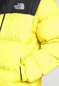 Żółta kurtka puchowa z czarnym panelem na ramieniu, zamek błyskawiczny i ocieplane rękawy. Na mankiecie znajduje się napis "700" oraz logo "THE NORTH FACE".