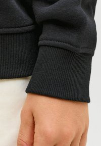 Puño de sudadera negra con textura acanalada y costuras impecables. La vista en primer plano muestra un tejido suave y un diseño ajustado para un uso ceñido.