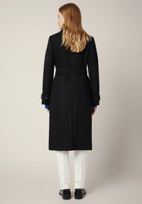 Czarny wełniany długi płaszcz z paskiem w talii, bocznymi kieszeniami i guzikami przy mankietach, noszony na białych spodniach i czarnych butach, widziany z tyłu.