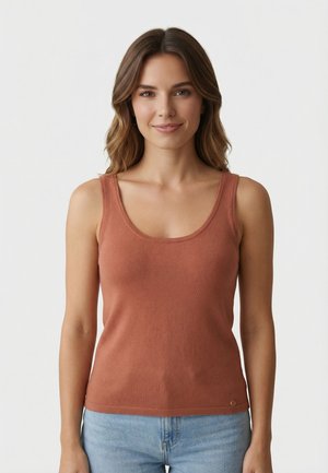 Jeune femme aux cheveux bruns ondulés portant un haut sans manches couleur rouille et un jean bleu clair, souriante sur un fond blanc uni.