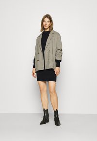 Dubbelknäppt blazer i houndstooth-mönster, beige och svart, med vadderade axlar. Bärs över en figurnära svart tröjdress och ankelskor.