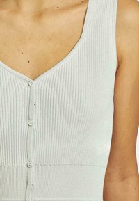 Top a coste grigio chiaro con scollatura a V, dotato di bottoni decorativi e design aderente. Il tessuto ha una leggera lucentezza e una consistenza morbida.