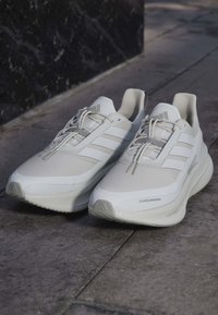 Pari valkoisia Adidas juoksukenkiä nauhoilla harmaalla kadulla lähellä mustaa seinää, sivu- ja etunäkymä.