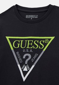Fekete organikus pamut póló kerek nyakkal. Zöld és szürke háromszög alakú hímzett logóval rendelkezik, rajta a következő felirattal: "GUESS U.S.A. WASHED JEANS."